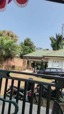 The video shows a view from a covered porch or balcony looking out towards a building and surrounding greenery. In the foreground, a black metal railing with vertical bars is visible. Behind the railing, several motorcycles are parked, some with colorful accents. A black SUV is parked in front of a low-rise building with a green tile roof. The building appears to have an entrance with a sign that reads "BADAN SERTIFIKASI INSINYUR INDONESIA". There is a cell tower visible in the background behind the building, amidst trees and foliage. The sky is clear and blue, indicating a sunny day. The overall scene suggests an outdoor area, possibly a parking lot or entrance to an office building, with a natural and somewhat rural setting. There are no people clearly visible performing actions, though some figures appear to be near the motorcycles and the building entrance, suggesting activity. The camera is static, capturing a wide view of the scene.