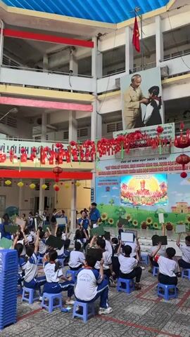 An outdoor school event is captured in a bright, multi-story school courtyard in Ho Chi Minh City, Vietnam. Numerous students, dressed in white shirts and dark blue uniforms, are seated on blue plastic stools, actively participating in an activity. Many students are raising their hands and holding up small white boards, indicating they are answering questions or engaging in a game.

At the front of the courtyard, an improvised stage or activity area is set up with a large projection screen. The screen displays colorful graphics and Vietnamese text, including "CHỦ ĐỀ NĂM HỌC 2023 - 2024" (Theme of the school year 2023 - 2024) and phrases related to a "Book Reading Festival" or "Reading Culture in School," along with images of books. Several adults, likely teachers or event organizers, are present, guiding and observing the students.

The courtyard is decorated with numerous red lanterns and banners featuring Vietnamese text. A large portrait of Ho Chi Minh is prominently displayed above the stage, with a Vietnamese flag flying on a pole above it. The event takes place during the daytime, under bright, clear skies. A stack of blue plastic stools is visible on the left side of the frame. The atmosphere appears lively and engaging, with students actively involved.