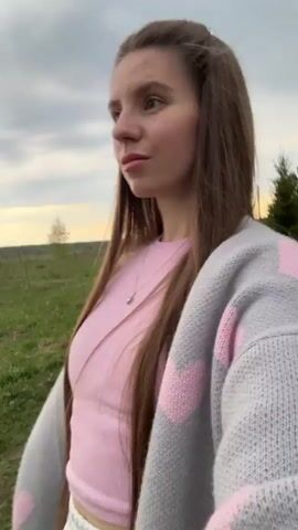 A woman with long brown hair, wearing a pink top and a gray sweater with pink hearts, walks on a grassy path while holding hands with a small child in a beige outfit and a red cap. The woman holds the camera, filming herself and the child. The background is a field under an overcast sky.