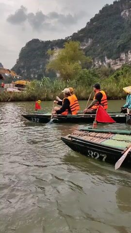 Several narrow boats are being rowed down a river. Each boat holds multiple passengers wearing orange life vests. The river is surrounded by green vegetation and mountains in the background. The sky is overcast. Some boats have small flags on them, and boat numbers are visible. Passengers are using oars to propel the boats. One person is wearing a conical Asian hat. An excavator is visible on the riverbank in the distance.