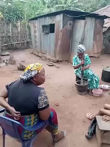 Several people are gathered in a dirt courtyard. One woman sits on a chair and speaks. Another woman sits on a stool and grinds something in a mortar with a pestle. Other women sit nearby. A child in a red outfit sits on a stool. A young woman walks by carrying a plate. The courtyard is surrounded by simple buildings, including a corrugated metal structure and a brick building with a small window. There are pots, buckets, and other household items scattered around. Some of the people are wearing traditional African clothing.