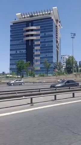 A white van drives past on a highway in front of a modern glass office building. Several other vehicles, including a large white truck and other cars, are visible on the highway. The sky is clear and blue.
