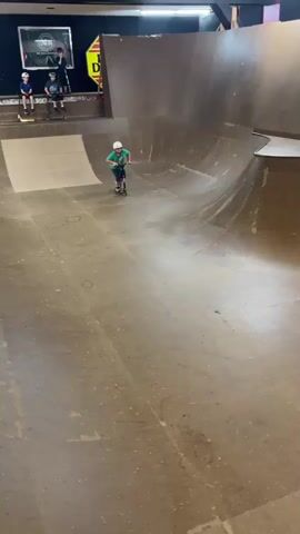 The video shows a young boy wearing a green t-shirt, blue shorts, and a white helmet riding a scooter in a skatepark. He is in a large bowl-shaped ramp area. The boy rides towards the camera and then turns. He appears to lose control and falls onto the ramp, with his scooter flying off to the side. In the background, there are other people watching from a platform above. The environment is a skatepark with wooden ramps.