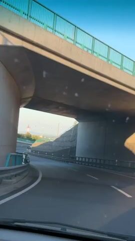 The video shows a first-person perspective of driving on a road that curves to the left under an overpass. The road is asphalt with white lane markings. On the right side of the road, there is a retaining wall made of gabions, with red and white striped markers along the guardrail. On the left side, there is another retaining wall with a green guardrail. Trees and a small building with a red roof can be seen in the distance to the left, under a clear blue sky. The sun appears to be low in the sky, casting shadows. The video is short, showing the car moving forward along the curved road.
