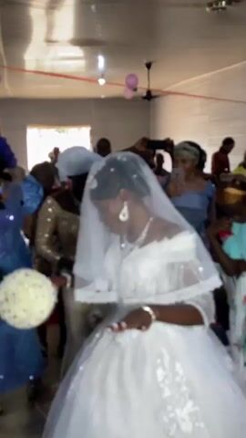 The video shows a bride in a white wedding dress, holding a bouquet, walking through a brightly lit room with a low, white ceiling. The room is decorated with pink balloons. Several women in formal attire are walking in the same direction, some of them taking pictures with their phones. The video is shaky and the focus is inconsistent, sometimes blurring the bride and other people in the scene. Overall, the setting appears to be a wedding celebration, likely taking place indoors.