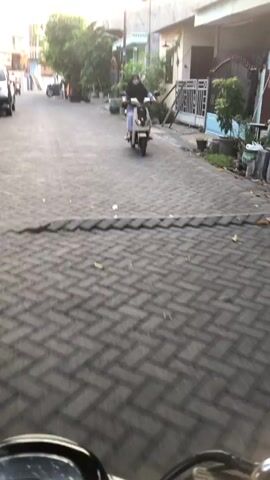 A person rides a white scooter down a paved street. The rider is wearing dark clothing and a mask. Trees and houses line the street. The sun appears to be setting, casting long shadows. The camera is mounted on another scooter, following the first one.