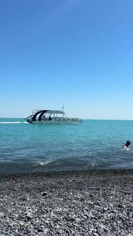 The video shows a boat moving across turquoise water under a clear blue sky. The boat is white with a black canopy and has the word "АЙСУЛТАН" and "АЛАКӨЛ" written on its side in dark lettering. A flag, likely the flag of Kazakhstan, flies from a pole on the boat. The boat appears to be a passenger vessel, possibly for tours. In the foreground, a pebble beach is visible with small waves lapping at the shore. A person is in the water near the shore, facing away from the camera, with their head and shoulders visible. The overall scene suggests a sunny day at a lake or sea.