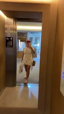 The video shows a woman standing in an elevator, taking a selfie in the mirrored doors. She is wearing a white t-shirt, white shorts, a pink baseball cap, and pink Crocs. A light-colored shirt is tied around her waist. She is also carrying a brown bag over her shoulder and a white bag in her left hand.

The elevator appears to be modern, with stainless steel doors and wooden paneling on the walls. The reflection in the mirror shows a hotel hallway with a carpeted floor, a spiral staircase, and some furniture. The lighting in the elevator is bright and artificial.

The woman seems to be posing for the photo, looking at her phone as she takes the selfie. Her facial expression is neutral. The overall impression is that of someone casually dressed and perhaps checking her appearance before heading out.

There is text visible on a sticker on the elevator door, but it is mostly illegible. It appears to be an elevator safety notice or a building directory.

The location is identified as Žunovnica, Bosnia and Herzegovina. The time of day and weather are not discernible from the video.