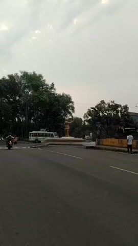 The video shows a wide, paved road in an urban setting. In the middle of the roundabout there is a bronze statue of a man standing with his arm raised. To the left of the statue is a large green and white bus. Motor scooters are moving on the road. A man walks on the road’s edge on the right. The sky is overcast.