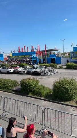 The video shows a monster truck show. The truck drives over a row of cars and a metal frame, crushing them in the process.  There is a crowd watching behind a metal barrier.  The sky is blue, and there are flags and banners in the background advertising the "World of Motorshow." Two people in the foreground are recording the event with their phones.