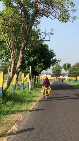 A person wearing a headscarf and a patterned dress walks along a paved road. They are carrying a bag. The road is bordered by a grassy area and a fence with yellow and blue posts. Trees line the left side of the road, and more trees and buildings are visible in the distance. The sky is clear and blue, and the sun is casting shadows. The scene appears to be in a rural or suburban area.