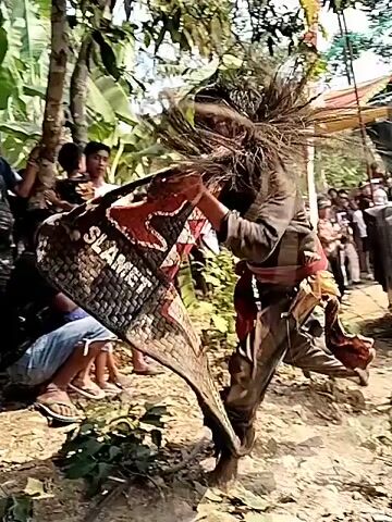 The video begins with a close-up of a person wearing a colorful, patterned garment, possibly a costume or traditional attire, with a blurred background of foliage and other people. The camera then pans to show a group of individuals, mostly women, standing in what appears to be an outdoor gathering or event. Some are wearing headscarves. The scene transitions to a wider shot of a crowded area, with more people visible, some of whom are children. There are structures resembling pillars and roofing, suggesting a covered outdoor space or a building entrance. The people in the video are facing towards the camera or interacting amongst themselves, with expressions ranging from neutral to smiles. The environment appears to be tropical or subtropical, with lush greenery visible in the background. The video captures a moment of community interaction, possibly a celebration or a public event.