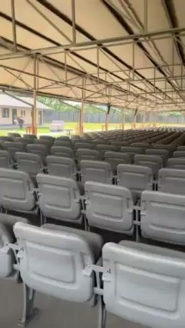 The video shows an outdoor auditorium with a metal roof structure. The auditorium is filled with rows of gray plastic chairs. There is a stage at the front of the auditorium.  A person can be seen walking among the rows of chairs.