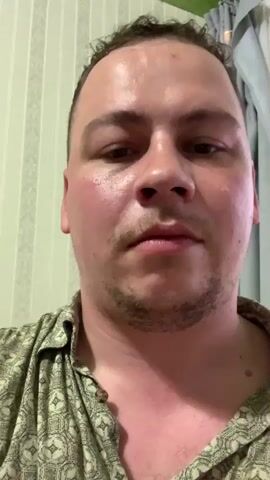A close-up, front-facing shot shows a man speaking. He is wearing a patterned collared shirt. His skin is flushed, and he appears to be sweating. The background consists of striped wallpaper and a curtain.
