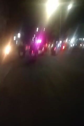 A blurry, night-time video shows a street scene with moving vehicles. The camera appears to be mounted on a moving vehicle. Multiple motorcycles and cars are visible, their headlights and taillights creating streaks of light. One motorcycle in the foreground is mostly visible. In the middle ground, there is a cluster of vehicles with flashing lights, appearing to be emergency vehicles. One individual is visible walking or standing near the emergency vehicles. The street is paved and appears to be illuminated by streetlights and vehicle lights. The overall impression is a busy street scene at night, possibly with an incident underway due to the flashing lights.
