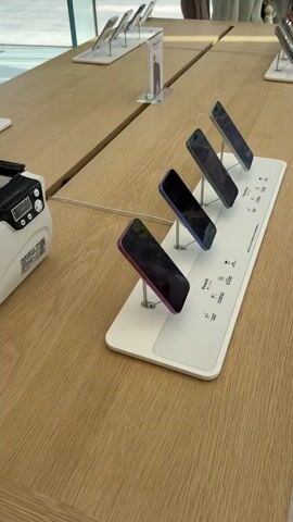 A white, rectangular, wood-grained table holds several Apple iPhones on display in an Apple store. Each iPhone is mounted on a small, individual, angled, metal stand and has a small sign next to it. A white and black bill counter is on the left side of the frame. A person wearing a striped shirt reaches for an iPhone on the table in the background, out of focus. The store has bright lighting and a neutral aesthetic. There are other customers in the background. The iPhones on the table are arranged linearly.