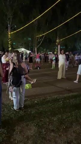 A group of people are gathered outdoors at night, possibly at an event or festival. They are standing on a dark paved area surrounded by grass and trees. Yellow string lights are visible above the crowd, illuminating the scene. Many individuals are dancing and interacting, with some wearing brightly colored clothing. In the foreground, a few people are partially visible, including one person sitting down. The overall atmosphere appears lively and festive.