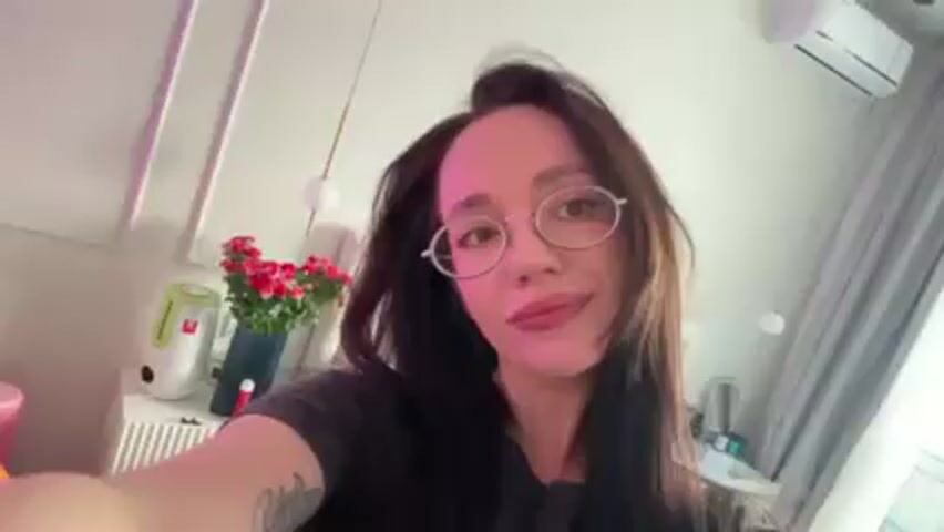 The video is a selfie taken by a woman with long dark hair and glasses, smiling slightly at the camera. She is wearing a dark grey shirt. The background includes a white wall with framed panels and a small vase of red flowers on a table to her left. Other objects on the table include a humidifier, a small figure, and a lamp.  Above the table, a hanging light fixture is visible. Behind her is a bed with blue bedding and a white headboard.  An air conditioning unit is mounted on the wall near the ceiling.  The lighting in the room is soft, and the woman's face is well-lit.  A tattoo is visible on her left arm.