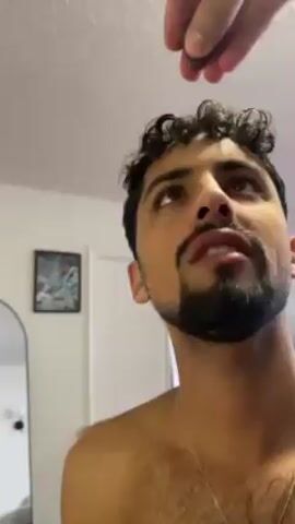 Here's a concise description of the video:

The video shows a man with curly hair and a beard tilted his head back with his mouth open. Another person's hand reaches into the man's mouth and appears to put something inside. The man then closes his mouth, smiles, and seems to swallow it. The background includes a white wall, a framed picture, and a mirror. The man is shirtless.