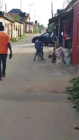 The video captures a daytime street scene in Ohokobe Ndume, Nigeria. In the foreground, a person in an orange shirt walks away from the camera on the left side of the paved road. Central to the frame, two young males are engaged in what appears to be a playful grapple or mock fight. One wears a blue shirt and the other a lighter-colored outfit.

A woman in a vibrant, patterned dress walks from the background towards the center, passing a parked black car. Another child, dressed in a light top and skirt, stands near the woman. On the right side of the street, there are small shops or stalls, displaying goods like clothing and several large sacks. Buildings line both sides of the street, some with visible water tanks on their roofs, and power poles are present. The sunlight suggests it is daytime. No visible text is present.
