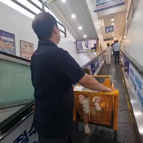 A man is pushing a shopping cart with a child in it up an escalator. The man is wearing a black shirt and the child is wearing a light-colored outfit. The escalator is moving upwards and there are other people on the escalator further up. The walls of the escalator have advertisements and signs. The lighting is bright, indicating an indoor environment.