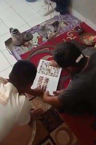 Two people, one wearing a white shirt with text and the other in a dark shirt, are looking at a book or magazine spread open on the floor. They are on a rug with a patterned design, and a tiled floor is visible. There is a kettle and other items on another rug in the background. The person in the white shirt is leaning closer to the book, touching it. The person in the dark shirt is also looking at the book, with their hand near it. The scene appears to be indoors.