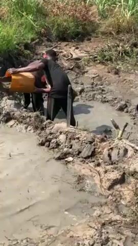 Two people are standing in a muddy, ditch-like depression. One person is on the left, wearing a dark shirt, dark pants, and a red cap. They are using a shovel. The other person is on the right, wearing a black t-shirt and dark pants. There's a metal post or stick partially buried in the mud near them. The ground around the muddy area is dirt and grass. The area is surrounded by tall green grass. Both people are digging and removing something from the muddy area.
