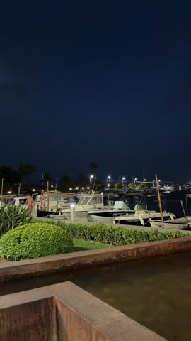 The video shows a night scene at a marina. In the foreground, a fountain sprays water with a warm light. Behind the fountain, several boats are docked. In the background, there are lights from a town or city and the silhouette of hills. The sky is dark blue with some clouds. There are a few people visible on the dock, walking or standing near the boats. The overall atmosphere is peaceful and serene.