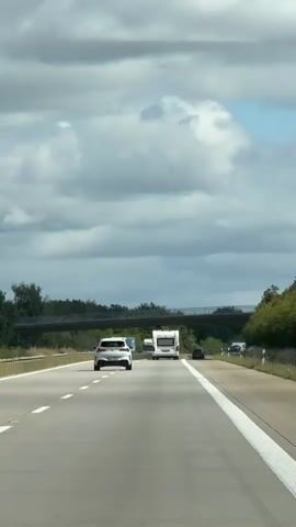 The video shows a view from a moving vehicle on a highway. The road has multiple lanes marked by white lines. In the foreground, the perspective is of the asphalt road surface with white lane markers. Further ahead, several vehicles are visible, including a white car in the left lane and a white camper van or RV in the right lane. Beyond these vehicles, a bridge or overpass spans across the highway. Trees and greenery line the sides of the road, and the sky above is partially cloudy. The overall scene depicts highway travel on an overcast day.