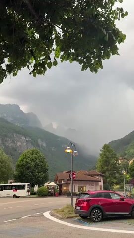 This vertical media file captures a scenic view of a town nestled at the base of majestic mountains. A lush green tree with vibrant leaves partially frames the top of the image, suggesting it is daytime, possibly late afternoon or early evening given the overcast sky.

The scene features a prominent red SUV parked in a designated parking area on the right side of the frame. In the mid-ground, a quaint building with a terracotta roof and arched windows stands, possibly a shop or restaurant, with a sign that appears to read "PIÙ BIO". A street lamp with a vintage design illuminates the area in front of it. To the left, a white bus is visible on the road, indicating potential public transportation or tourism.

The backdrop is dominated by imposing, verdant mountains, shrouded in a soft, misty haze, suggesting recent or ongoing rain. The weather appears to be cloudy and somewhat dramatic, with the fog lending an atmospheric quality to the landscape. The overall emotion evoked is one of serene natural beauty, with a touch of moody weather.

Visible text includes the word "STOP" on a red circular traffic sign and the partial word "PIÙ BIO" on the building. The location appears to be Molveno, Italy, known for its stunning alpine scenery. The presence of parked cars, a bus, and a building suggests a populated area within this natural setting. The grass and trees are lush, indicating a temperate climate.