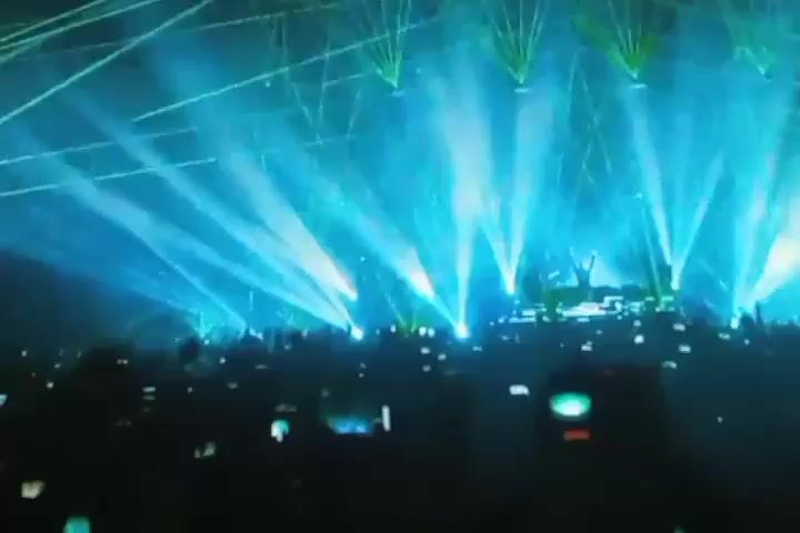 The video depicts a concert or large event. In the foreground, several people are visible, with hands raised in the air, some holding up cell phones and cameras. One person in the immediate foreground is holding a camera, and another is holding a cell phone. The background is dominated by bright lights, possibly stage lighting, and a hazy atmosphere, suggesting smoke or fog. The overall impression is of an energetic crowd at a performance venue.