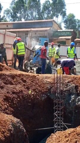 A video shows a construction site. A group of workers, some wearing safety vests and hard hats, are mixing cement with a cement mixer near a pit in the ground. The pit has steel reinforcement structures in it. The ground around the pit is dirt. One worker is using a board to move around the pit.