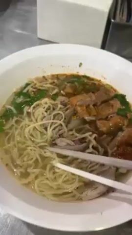 The video shows a bowl of noodle soup on a stainless steel surface. The soup includes noodles, pieces of meat, and green herbs. A pair of white chopsticks lifts a portion of noodles from the bowl. A white tissue box and a metal container are visible in the background.