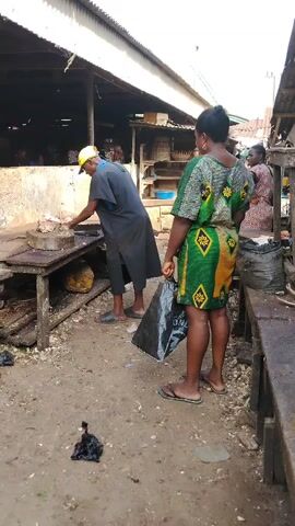 This image captures a vibrant, open-air market scene in Awka Etiti, Nigeria, likely during the daytime under clear weather conditions.

In the foreground and center, a man, appearing to be a butcher, is actively chopping what looks like meat on a round, wooden chopping block. He wears a dark, loose-fitting short-sleeved top or tunic, dark cropped trousers, and flip-flops. A bright yellow baseball cap covers his head. He holds a large, heavy cleaver, positioned as if mid-stroke or preparing to chop.

To the right of the butcher, a woman stands facing him, appearing to be a customer. She wears a striking knee-length dress with a geometric pattern in shades of green, yellow, and brown, and flip-flops. She holds a black plastic shopping bag in her left hand, which has partially visible white text, including the word "ONLY". She wears gold-colored hoop earrings. Her posture suggests she is waiting for her purchase.

The immediate surroundings include a rustic wooden table or stall where the butcher is working. The ground is unpaved, consisting of packed dirt, with some scattered debris and small black plastic bags visible.

In the mid-background, other individuals can be seen. One woman wears a light-colored headscarf and a green-and-blue patterned top. Further back, another woman is dressed in a reddish patterned garment. These individuals, along with other blurred figures, suggest a bustling market environment.

The market stalls feature simple wooden constructions and corrugated metal roofing, characteristic of many outdoor markets in the region. To the far right, part of another wooden stall or bench is visible, with dark sacks or bags placed on or near it. The overall setting conveys a sense of everyday commerce and community activity.