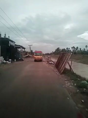 The video shows a wet road in a somewhat rural area. On the left side of the road, there's a building under construction and a few parked motorcycles. On the right, a riverbank is cluttered with trash. A yellow and red truck approaches in the distance on the road, moving towards the camera. The sky is overcast with gray clouds. Power lines run above the road.