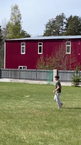 A young man is walking barefoot across a grassy lawn. The lawn is surrounded by a fence, behind which is a large, red building with white-framed windows. In the background, there are trees and a blue sky. The man is wearing a green t-shirt and light gray pants. He walks towards the camera.