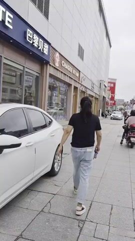 The video pans across a street scene in a city. In the foreground, a person with dark hair pulled back in a ponytail walks away from the camera, wearing a black t-shirt and light-colored jeans, holding a folded umbrella in their right hand. To the right, a young person with blonde hair tied in a ponytail walks away from the camera, wearing a white t-shirt and patterned shorts, carrying a silver backpack. Behind them, a person with dark curly hair walks away, wearing a green t-shirt and white shorts, carrying a brown bag. A scooter with a red storage box on the back is parked on the sidewalk. In the background, there are cars parked along the street, including a white sedan. Buildings line the street, with shopfronts and signage. A person is riding a scooter down the street. The sky is overcast.