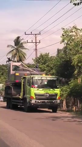 Here is a description of the video:

The video shows a green truck driving down a road. The truck is carrying an excavator with the words "ZE215E" on it. The truck has the name "LANCAR JAYA TRANSPORT" printed on its windshield. The road is paved and has white lines painted on it. There are trees and buildings on either side of the road. The video appears to be taken from inside a vehicle.