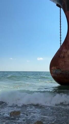 A video of a turbulent sea shore. In the foreground, a large rusted metal object, possibly part of a ship or buoy, is partially submerged in the water. It has graffiti on its surface. Waves crash against the shore, revealing pebbles and scattered driftwood. The water is a murky blue-green, with white foam from the breaking waves. In the mid-ground, a rope or cable stretches across the water, partially submerged and taut. In the background, the vast blue ocean stretches to the horizon under a clear blue sky with a few scattered white clouds. The sky is mostly empty, with no signs of human activity other than the distant presence of a large ship on the horizon. The overall impression is one of a wild and untamed natural environment, with man-made elements in a state of decay or abandonment.
