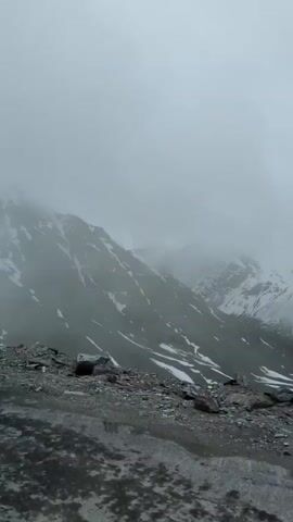 The video pans across a mountainous, desolate landscape shrouded in heavy fog and clouds. The scene is dominated by stark, rocky slopes interspersed with patches of snow, indicating a high-altitude, cold environment. The terrain appears rough and undeveloped, with loose rocks and gravel visible in the foreground. The weather is overcast and likely cold, with the thick fog obscuring the tops of the mountains and creating a sense of mystery and isolation. There are no people or visible objects in the frame, emphasizing the raw, untamed nature of the location. The overall mood is one of stark beauty and remoteness, characteristic of a harsh, high-altitude environment. The time of day is not discernible due to the atmospheric conditions. The location is identified as Zanskar Sumdo, India, which is a remote area known for its challenging terrain and weather. There is no visible text within the video.