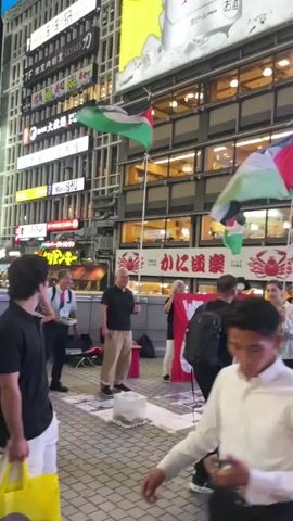 The video shows a street scene at night, with a group of people gathered. Several individuals are holding flags that appear to be Palestinian flags, which consist of three horizontal stripes of black, white, and green, with a red triangle on the hoist side. Some of the flags are attached to poles and are being waved or held aloft. In the center of the group, there is a red banner with white text that reads "WE STAND". Some of the individuals are looking towards the camera, while others are interacting with each other. The background features a tall building with illuminated signage, suggesting an urban commercial area. The ground is paved, and there are some objects scattered on it, including what appears to be a white bin. The lighting is primarily from the surrounding signs and ambient city lights. Some individuals are wearing casual clothing, while others are dressed more formally. One person in the foreground is partially obscured and appears to be wearing a white shirt. The overall atmosphere suggests a public gathering or demonstration.