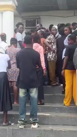 The video depicts a crowd of people of various ages and genders gathered outside a building with a light-colored exterior. Some individuals are sitting on the steps leading up to the building, while others are standing. The clothing styles vary, including casual wear, skirts, jeans, and vests. Some people are holding papers. The environment includes trees and a cloudy sky, suggesting an outdoor setting. The interactions between the people are not clearly discernible from the video, but it seems to be a gathering of some kind.