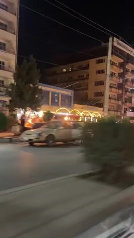 The video shows a nighttime street scene from a moving vehicle. The street is lined with buildings, some of which are illuminated with lights. There are cars on the street, and some people are visible. One building in the background has a sign that reads "Malwand University" in English. The camera is shaking, indicating that the vehicle is in motion. The overall impression is of a busy urban environment at night.