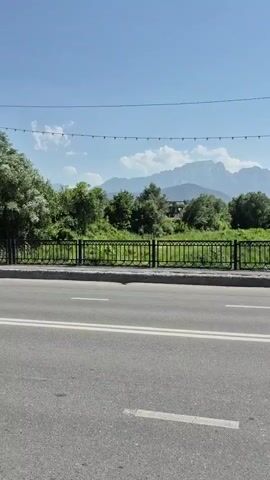 The video shows a view from a moving vehicle on a paved road. In the foreground, there is a gray asphalt road with white lane markings. A black metal fence runs along the side of the road, separating it from a grassy embankment. Beyond the fence, trees and vegetation are visible, with a body of water, likely a river, flowing in the background. In the distance, mountains can be seen under a clear blue sky with some white clouds. A string of lights is visible across the top of the frame. The camera appears to be panning slightly to the right as the vehicle moves.