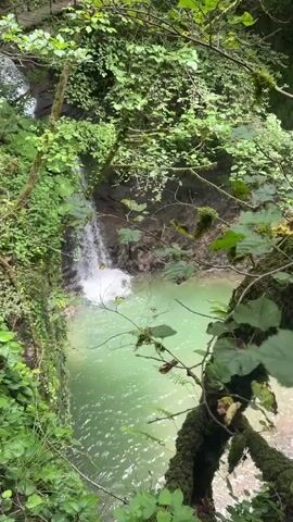 A short video shows a waterfall cascading into a turquoise pool of water surrounded by lush green foliage. The camera appears to be looking down at the scene from a slightly elevated vantage point. The waterfall is a thin stream of white water against a dark, rocky backdrop. The water in the pool is clear and reflects the surrounding greenery. The video is shot in landscape orientation and appears to be taken with a mobile phone. There is no audio accompanying the video.