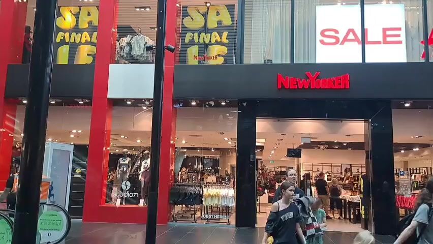 The video shows the exterior of a New Yorker clothing store. There are large windows displaying clothing and sales signs. In front of the store, several people are walking. The video is filmed from a slightly elevated position looking down at the store entrance. The lighting suggests it is daytime.