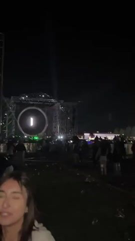 The video shows an outdoor concert at night. A large stage structure with a light-up circular screen is visible in the background, with flashing spotlights. In the foreground, two young adults, a man and a woman, are visible, partly obscured. The camera then pans away from the stage, revealing a panoramic view of a city skyline with twinkling lights in the distance. People are moving around, but the overall setting suggests a relaxed and celebratory atmosphere.