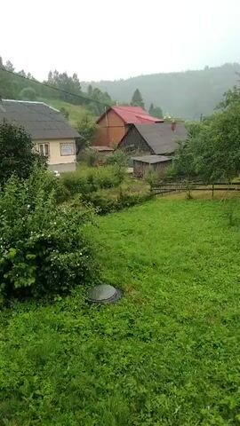 This video shows a rural scene with houses and lush green vegetation. A stream of water is visible in the foreground, appearing to be raining or a sprinkler system at work. Several houses with pitched roofs are scattered across the landscape, some with wooden siding and others with lighter colored walls. The houses are nestled among trees and rolling hills in the background, with a cloudy sky overhead, suggesting overcast weather. The overall atmosphere is one of a peaceful, natural environment.