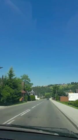 The video shows a POV shot from inside a car driving on a paved road. The road is lined with trees and houses on either side. The sky is clear and blue. The car is moving forward, and the scenery is passing by. There are a few other cars visible on the road. The overall impression is one of a scenic drive through a rural area on a sunny day.