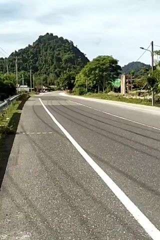 The video shows a man riding a scooter on an asphalt road with a solid white line dividing the two lanes of traffic. There are tire marks on the road. The man is wearing a grey jacket, black pants, and a white helmet. The road is surrounded by trees, grass, and a green mountain. There are also utility poles along the roadside.  The sky is cloudy.